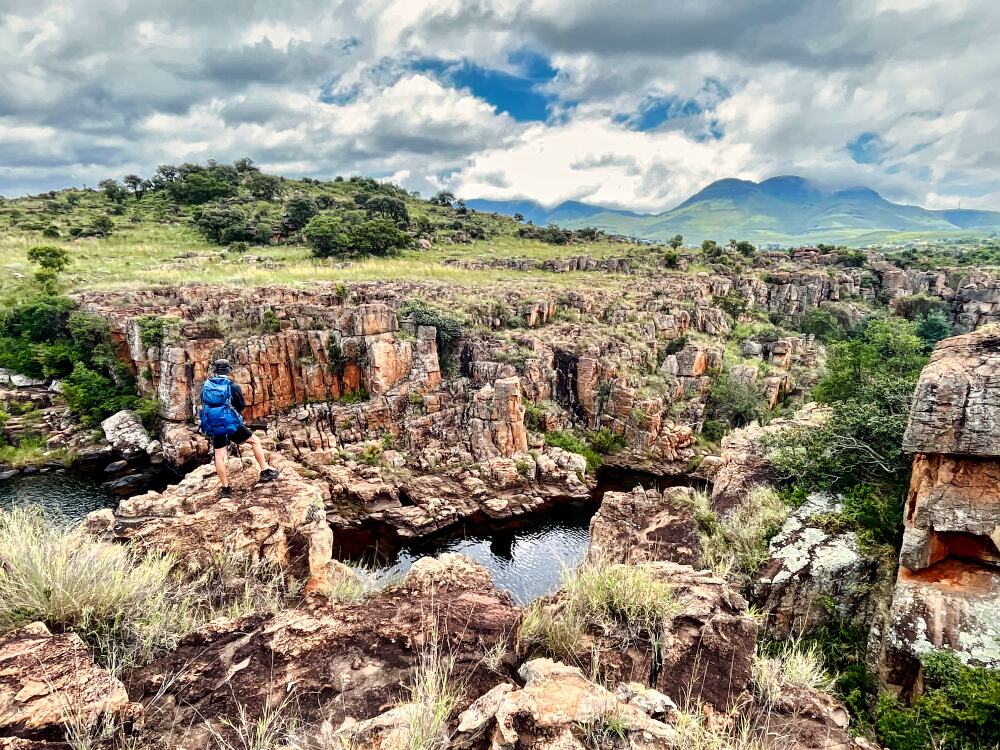 A hiker with a blue backpack stands on a rocky outcrop on the Blyde River Canyon hike with rugged, orange-tinted cliffs and a river below. The landscape is lush with greenery, and mountains are visible in the distance under a partly cloudy sky.